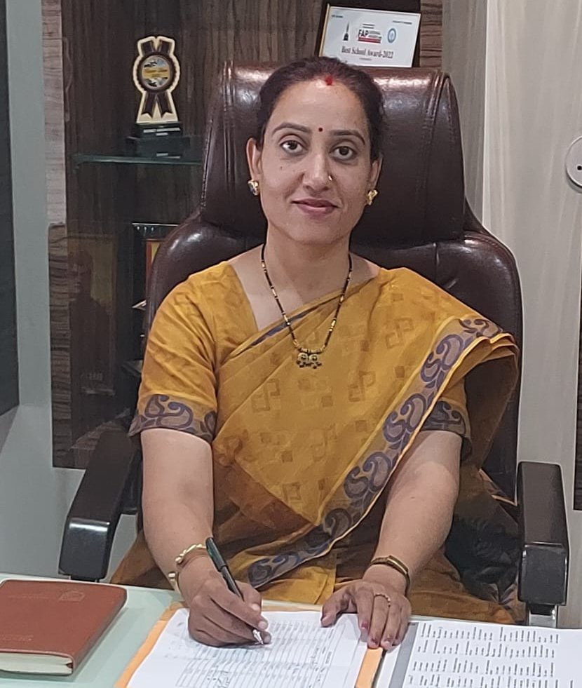 Woman in a mustard-yellow sari sits at a desk, signing a document with a pen while facing the camera in an office setting. Behind her are awards and a certificate on display.